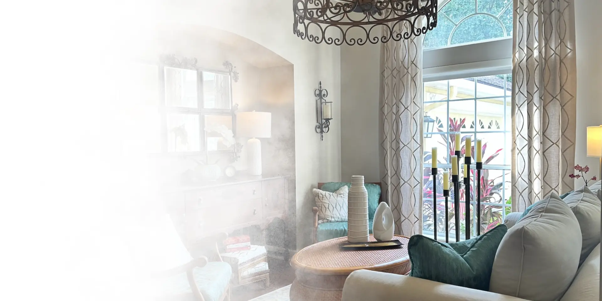 Sunlit Southern sitting room with an iron scroll chandelier, arched window, teal velvet pillows and a round rattan table