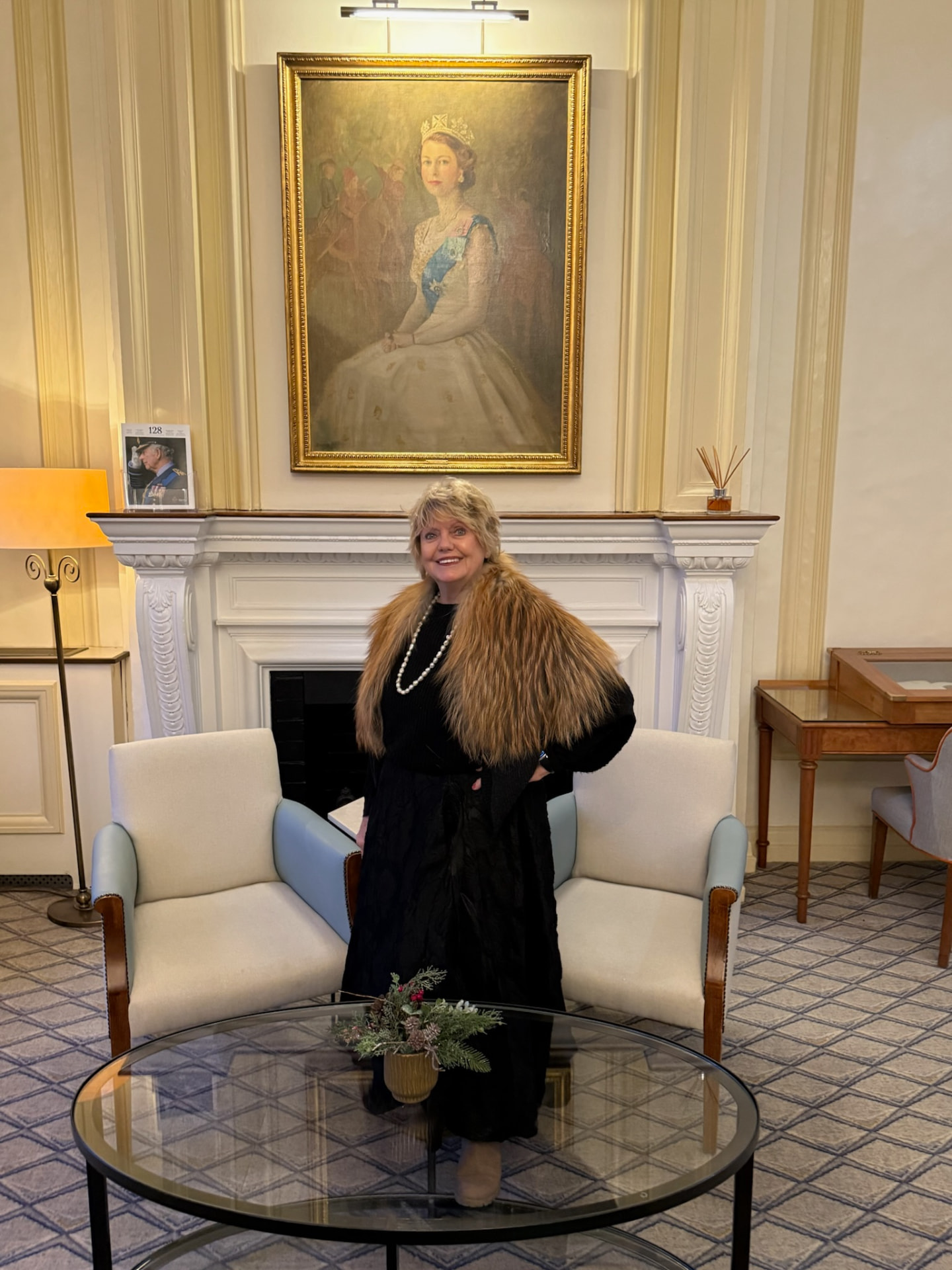 Madeline standing before a white marble fireplace and gilded portrait in an elegant panelled drawing room